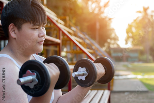 Young asian plump boy doing exercise with lifting heavy dumbbells in outdoor park in the sunset time of the day, sunlight edited.