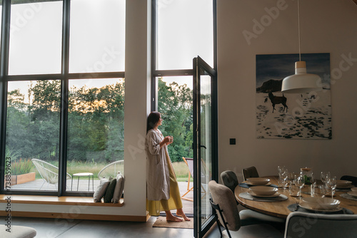 Woman standing with cup by the fireplace near the window at modern house on nature during summer time. Concept of vacation mood and comfort at home. Idea of recreation in cabins on mountains