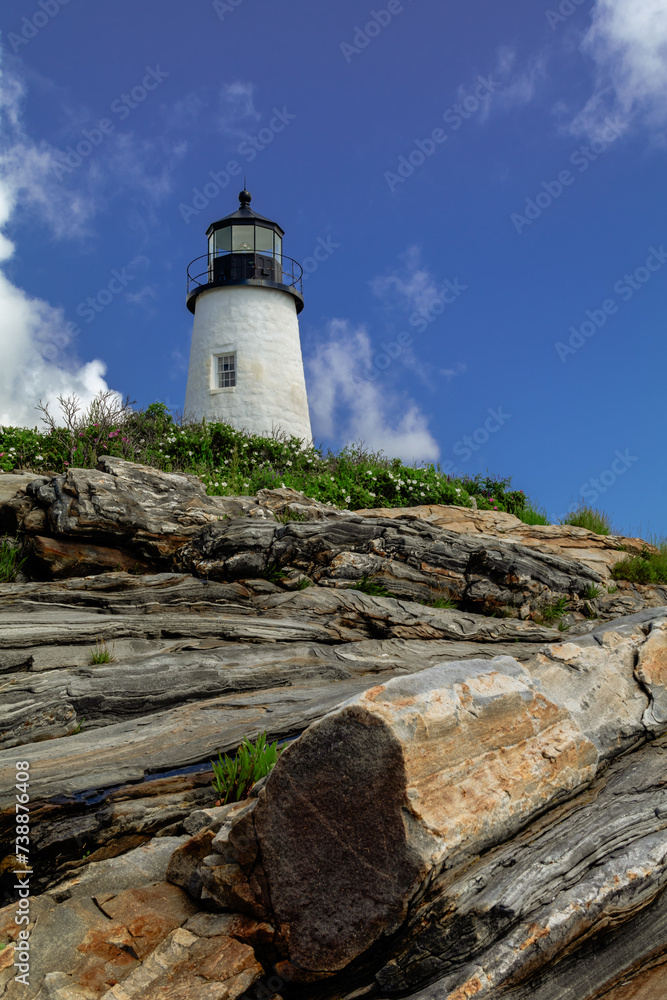 Pemaquid Point Lighthouse in Bristol Maine sits atop rugged rock layers on a bright summer day dotted with puffy clouds