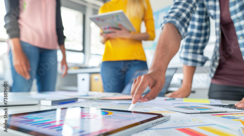 close-up of a person's hand holding a digital stylus over a digital tablet surrounded by colorful data charts, with other individuals collaborating in the background.
