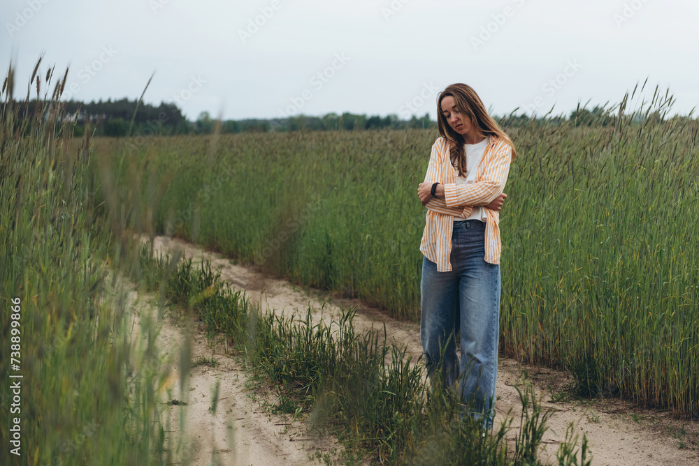 A girl, a woman stands on the road in the middle of a field of green wheat. Cool and cloudy summer day. He dreams with his eyes closed and his arms wrapped around himself A girl, a woman stands on the road in the middle of a field of green wheat. Cool and cloudy summer day. He dreams with his eyes closed and his arms wrapped around himself