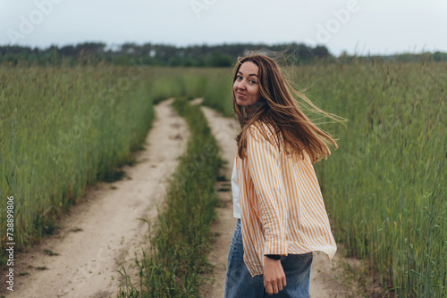 A girl, a woman walks into the distance along the road among a field of green wheat. Summer cloudy day. Hair blows in the wind. The girl turned around