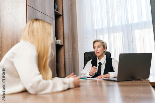View from table to professional middle-aged female teacher, manager or mentor helping young blonde student, new employee, teaching intern, explaining job, having teamwork discussion in office