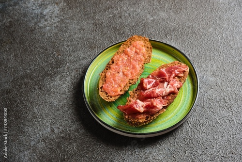 Spanish breakfast, two open tostada sandwiches with grated tomato and salted thinly sliced pork on dark bread on a green ceramic plate on a dark concrete background.