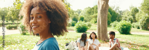 Portrait of young smiling woman with curly hair wearing blue t-shirt posing for the camera in the park, Panorama. Picnic on summer day outdoors her friends sitting in distance blurred on background