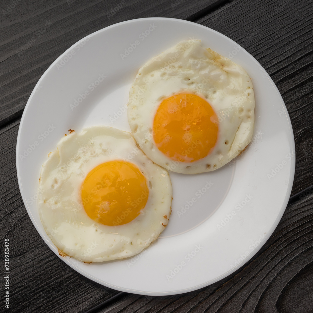 A white plate with two fried eggs and a sausage on a dark wooden surface. A white plate with two fried eggs and a sausage on a dark wooden surface.