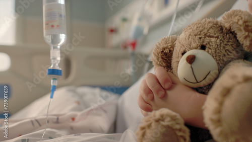 Close-up of young child's hand clutching a bear doll on bed in pediatrics ward with an intravenous drip visible in the background