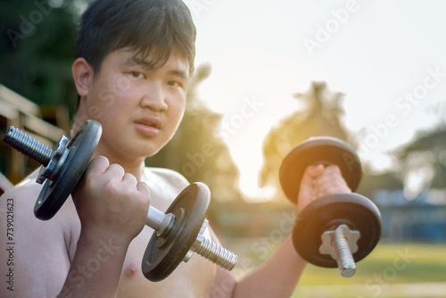 Young asian plump boy doing exercise with lifting heavy dumbbells in outdoor park in the sunset time of the day, sunlight edited.