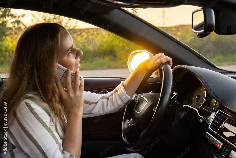 Naklejka premium Happy young woman speaking by mobile phone while driving car. Business woman talking phone call in automobile. Unsafely risky driving. Concept of multitasking 
