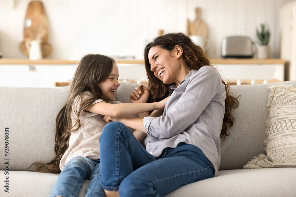 Beautiful Hispanic mother tickling cute little daughter sit on sofa, play on weekend, have fun, enjoy leisure and active game, feeling love, showing devotion. Family ties, playtime at home, motherhood