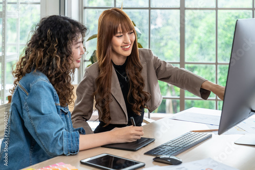 Two smiling young Asian female woman businessperson graphic designer working together, meeting and brainstorming, using computer in office, pointing at screen.