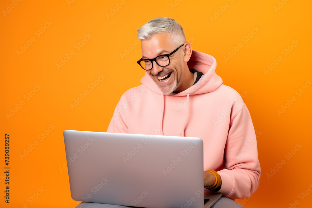 Joyful Man with Silver Hair Using Laptop on Vibrant Orange Background. Modern Remote Working Concept Joyful Man with Silver Hair Using Laptop on Vibrant Orange Background. Modern Remote Working Concept