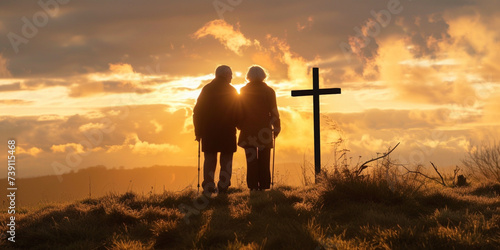 Silhouette of senior couple on high hill with cross during sunrise or sunset. Regretting sins, missing people who passed away, deeply religious person, praying, thinking about soul and meaning of life photo