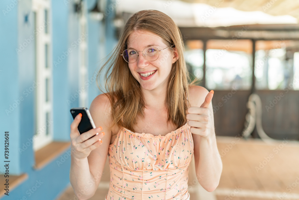 Young French girl with glasses at outdoors using mobile phone while doing thumbs up