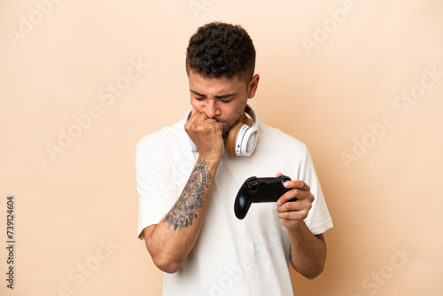 Young Brazilian man playing with a video game controller isolated on beige background having doubts © luismolinero