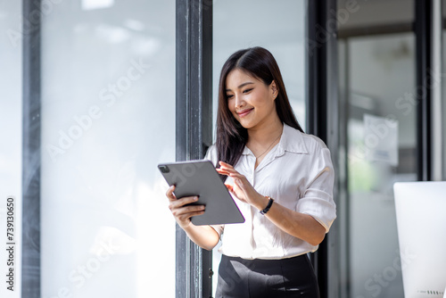 Young asian busy professional business woman executive, female company worker or manager holding digital tablet technology device working standing in modern corporate office.