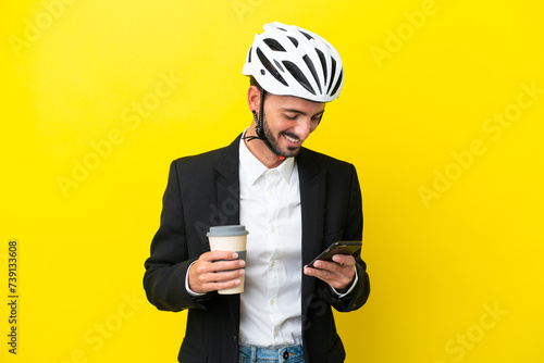 Business caucasian man with a bike helmet isolated on yellow background holding coffee to take away and a mobile