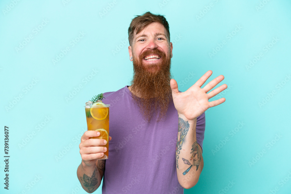 Redhead man with long beard holding a cocktail isolated on blue background saluting with hand with happy expression Redhead man with long beard holding a cocktail isolated on blue background saluting with hand with happy expression