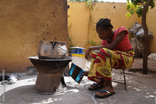Young African girl sitting in front of a simple charcoal stove fanning the fire; traditional food preparation