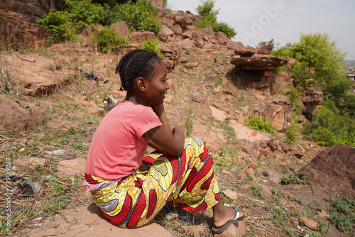 Side view of a serene daydreaming African villagegirl sitting on a rock amidst weeds and brushwood on a sunny day symbolizing differences in lifestyle and youth in developing country