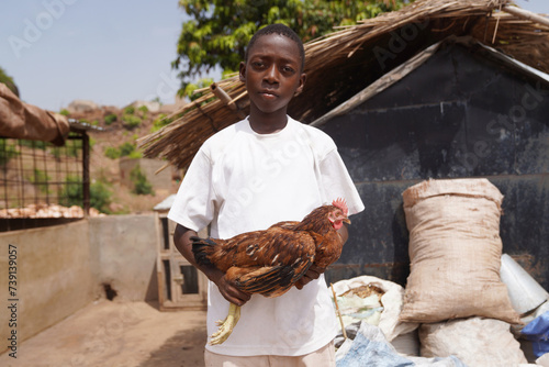 Pensive African farmboy with rooster standing in front of bags of chicken feed supply - traditional poultry farm scene