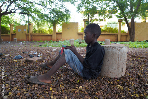 A lone black street urchin sitting on the ground with his begging pot staring into space in a run down African city setting