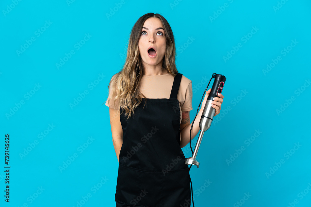 Romanian woman using hand blender isolated on blue background looking up and with surprised expression