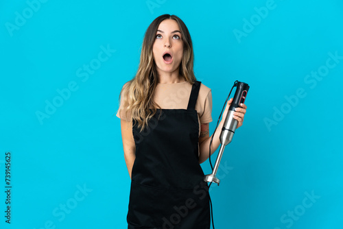 Romanian woman using hand blender isolated on blue background looking up and with surprised expression