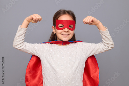 Strong powerful little girl wearing superhero costume and mask isolated over gray background standing with raised arms showing her biceps looking smiling at camera