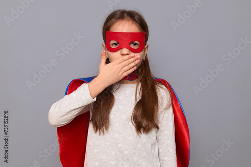 Shocked surprised little girl wearing superhero costume and mask isolated over gray background standing looking with surprised eyes covering mouth with palm sees something amazing