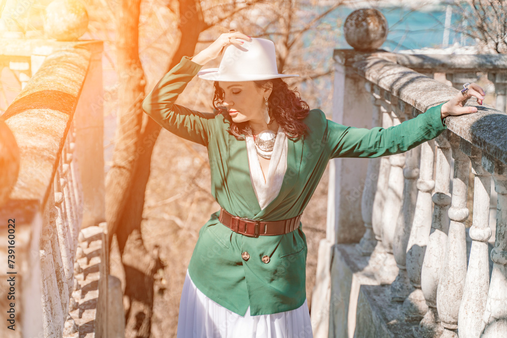 Woman walks around the city, lifestyle. Happy woman in a green jacket, white skirt and hat is sitting on a white fence with balusters overlooking the sea bay and the city.
