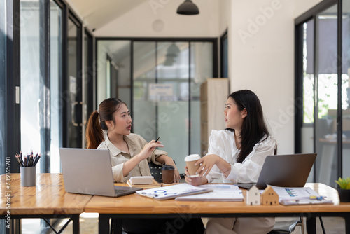 Two female coworkers are relaxing with coffee with laptops and documents on a table in a bright office.