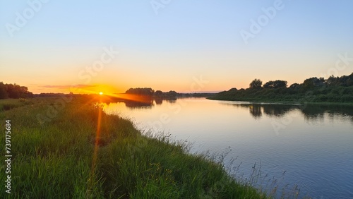 On a summer evening  the sun dips below the horizon and paints the sky in bright colors. There is a light ripple on the water. Tall grass and deciduous trees grow on the banks and reflect in the water