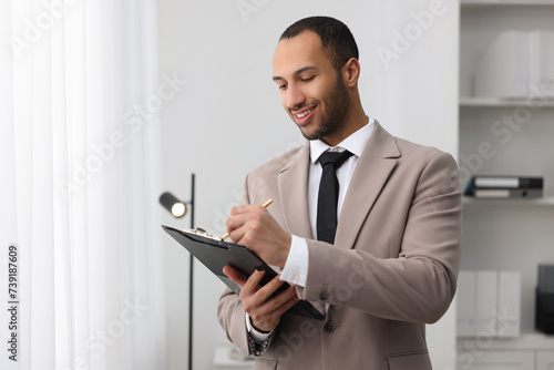 Smiling young man with clipboard writing notes in office. Lawyer, businessman, accountant or manager