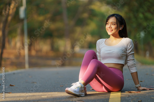 An Asian woman sits on the sidewalk, enjoying a well-deserved rest after a run in the park. © Songsak C