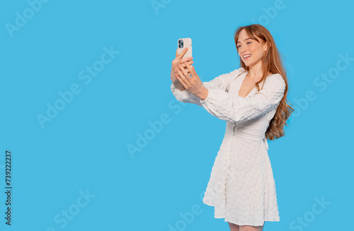 young redhead positive cute attractive woman in white dress holding a mobile cell phone chatting on a blue color background studio portrait