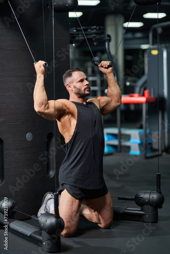 Serious sportsman wearing black tank top, doing exercise for biceps, standing on knees. Side view of muscular young man pulling cables of training apparatus, keeping muscles in tense. Sport concept.