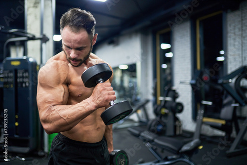 Fit topless man exercising in gym. Side view of strong bearded bodybuilder working out with heavy dumbbell weights, looking at bicep, against blurred background. Concept of sport, bodybuilding.