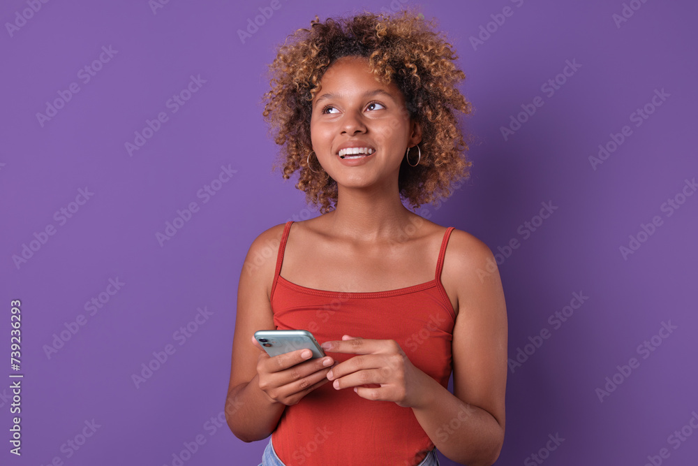 Young cute dreamy African American woman teenager uses smartphone and looks up figuring out what to buy in mobile online store application for shopping stands posing on purple background. Young cute dreamy African American woman teenager uses smartphone and looks up figuring out what to buy in mobile online store application for shopping stands posing on purple background.