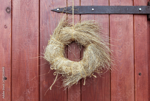 Wooden, red door with an old metal hinge. A round wreath of dry grass, tied with hemp rope, hangs on a rusty nail. Design.
