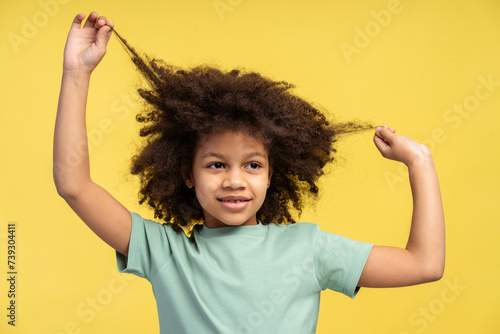 Happy African American kid elevates her arms, yanking her locs upwards, isolated yellow background photo