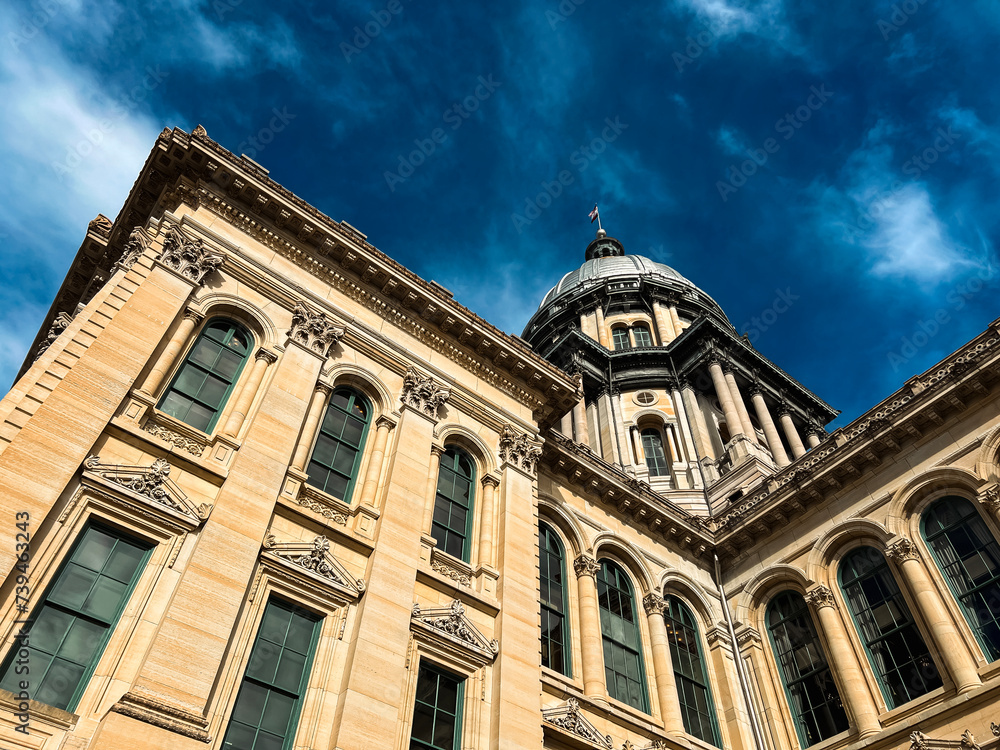 Fototapeta premium Rear views of the Illinois State Capitol Building in Springfield, Illinois, USA. Cloudy blue skies overhead. The US flag, State of Illinois Flag, and Black History Month Flag fly atop the dome.