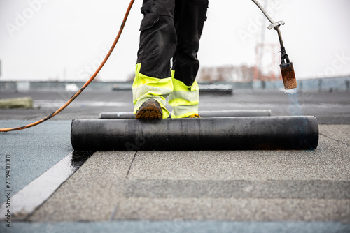 professional roofer applying tar with blowtorch on new roofing felt for waterproofing a flat roof construction site with safety gear photo