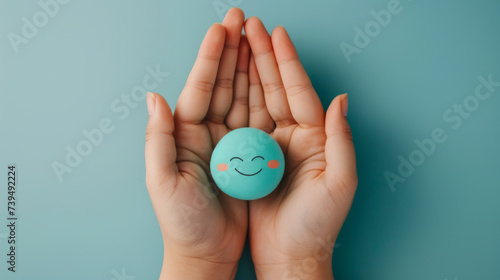 Two hands gently hold a smiling blue sphere against a calming turquoise background, conveying a sense of joy and emotional well-being.