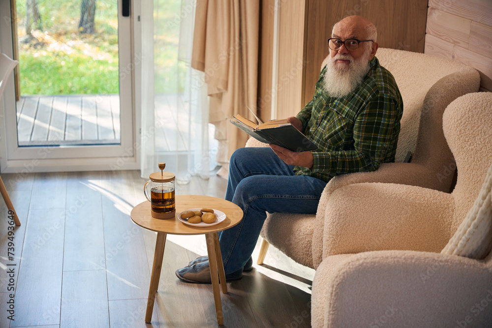 Gray-bearded old man in glasses settled down with book by French window