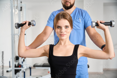 Female performs exercises with dumbbells under supervision of physiotherapist