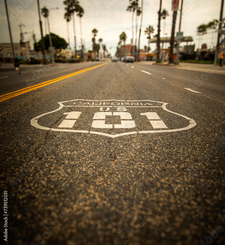 Highway 101 sign painted on the black asphalt road with city diffused in the background. The shot is from Oceanside California