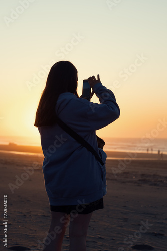 Mujer tomando una fotografía con su teléfono móvil en la playa durante el atardecer photo