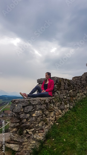 Woman sitting on medieval stone walls. Scenic view of castle ruins Burgruine Griffen in Voelkermarkt, Carinthia, Austria. Tourist attraction on cloudy day. Historic travel destination photo