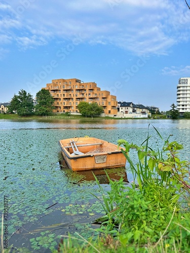 duckweed on the river, small boat on the water, buildings in the background, green leaves on the water